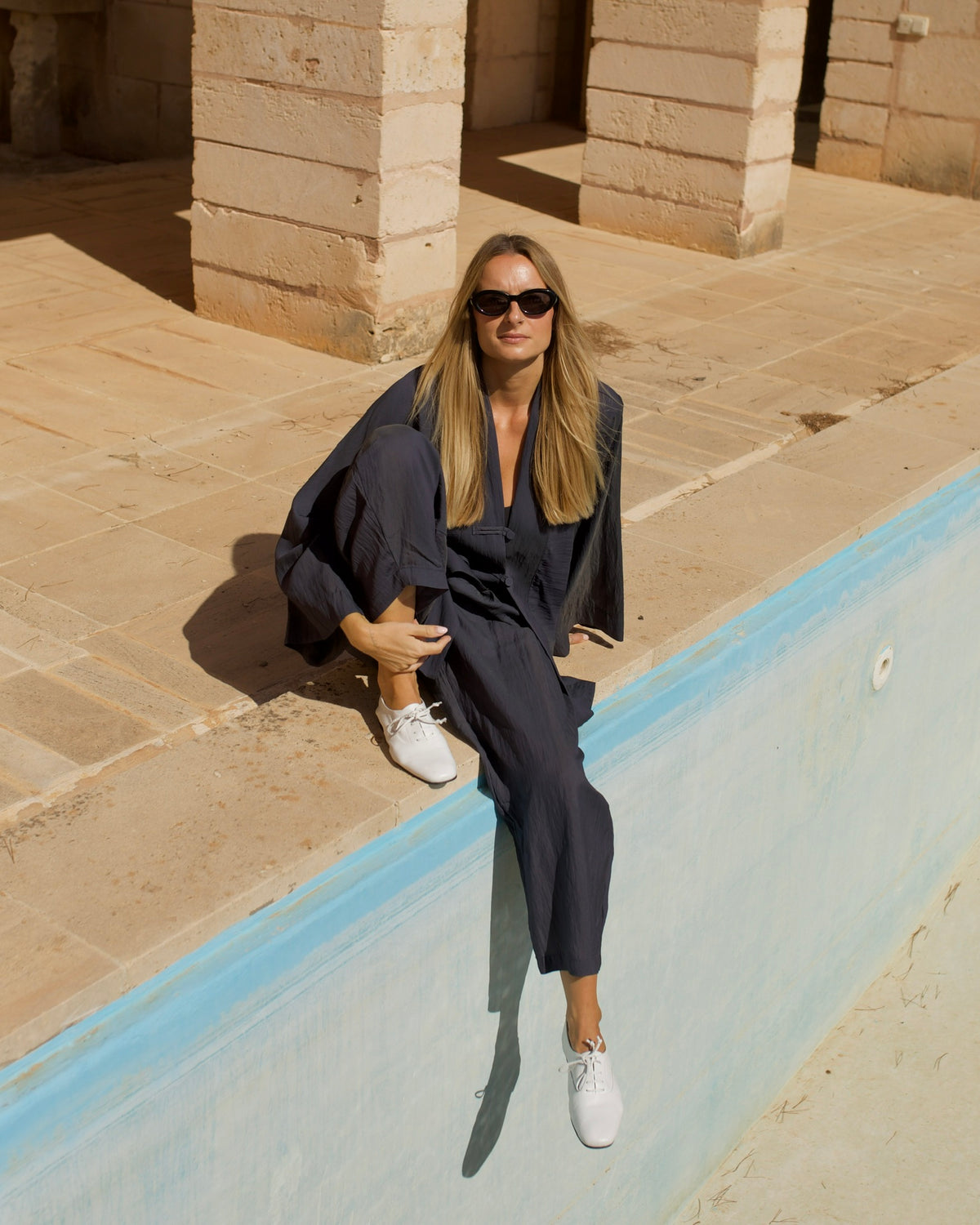 Woman in a navy linen set on the edge of an empty pool, wearing white Inca Oxfords