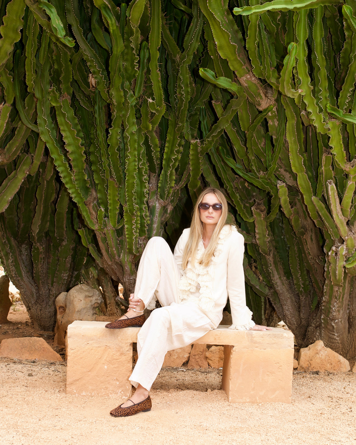 Woman in a white ruffled jacket on a stone bench under tall cacti, wearing cheetah-print Deia ballet flats