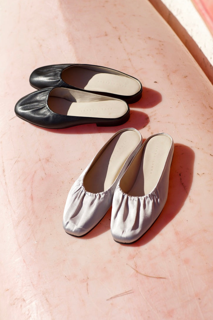 Arta ruched mules in black leather and grey satin on a sunlit pink surface