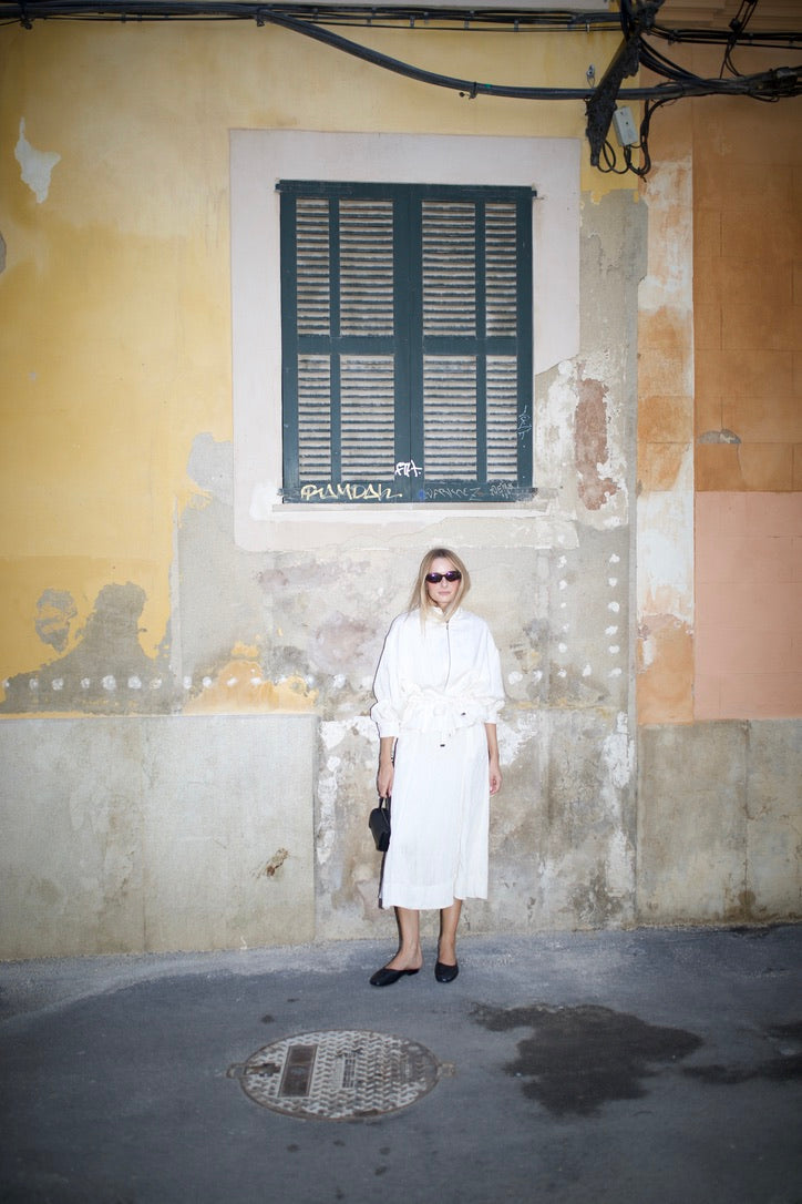 Woman in a white dress by a weathered yellow wall with green shutters, wearing black ballet flats