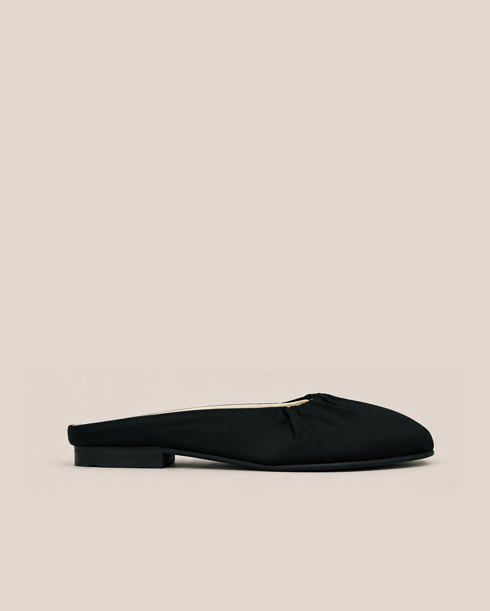 A single black satin mule flat shoe with a low block heel, shown in profile against a solid light beige background.
