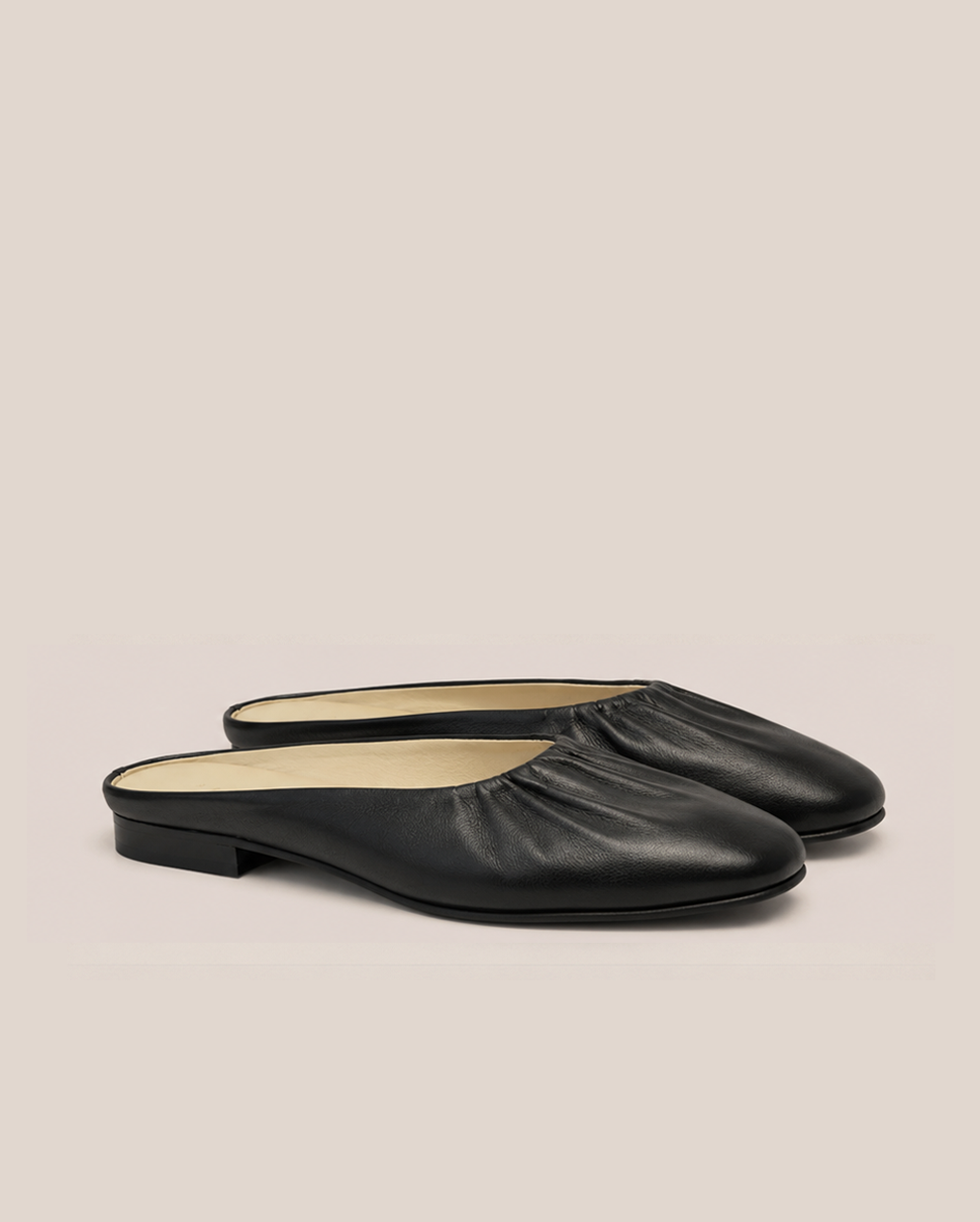 A pair of black leather slip-on mule flats with a gathered elastic top and low block heel, shown on a solid light beige background.
