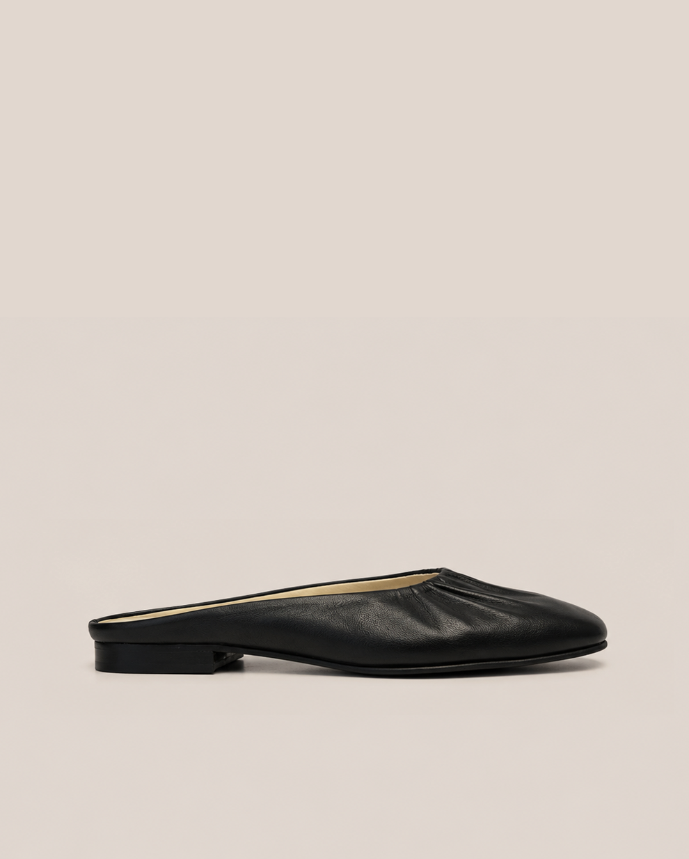 A single black leather flat mule with a pointed toe and gathered upper, shown in profile against a solid beige background.