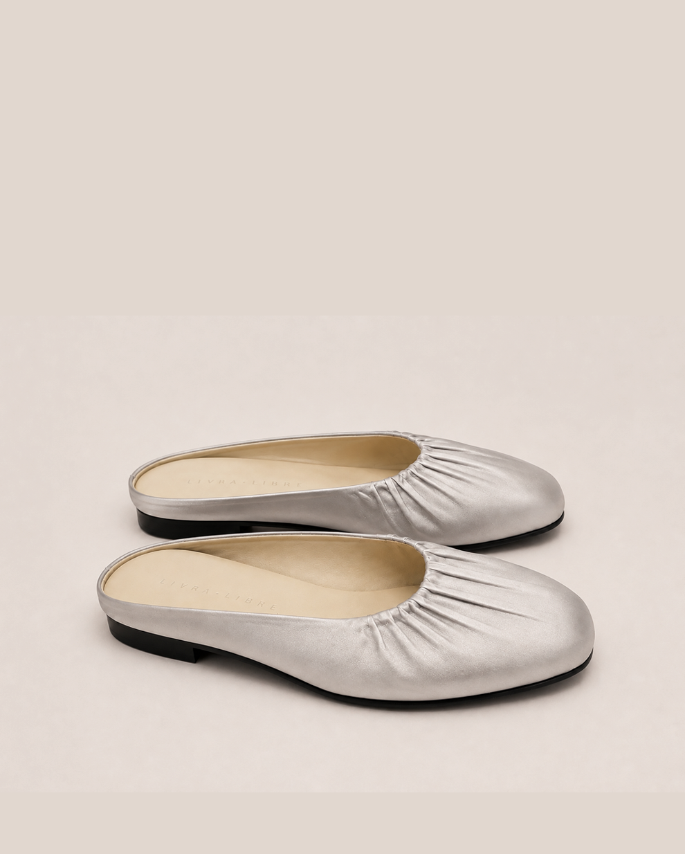 A pair of metallic silver flat mule shoes with gathered detailing around the square toes, shown on a neutral beige background.