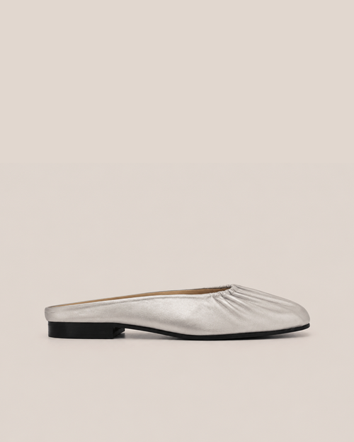 A single metallic silver flat mule slipper with a gathered, pleated crown and a low black heel, shown in profile against a solid beige background.